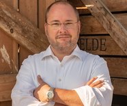 Porträt von Hauke Rüsbüldt, Geschäftsführer des Guts Wulksfelde im weißen Hemd mit verschränkten Armen vor einer Holzwand