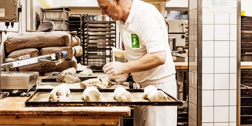 Bäcker formt Backwaren auf Arbeitsfläche, davor ein Backblech mit geformten Broten, im Hintergrund Backstube mit Papierzutatensäcken, Rollwagen mit Backblechen und Öfen