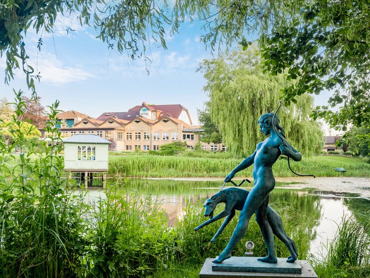 Im Hintergrund ein Gebäudekomplex aus Restaurant und Hotel, im Zentrum ein Gewässer im Grünen, im Vordergrund eine Statue eine Frau mit Bogen mit Hund an der Leine